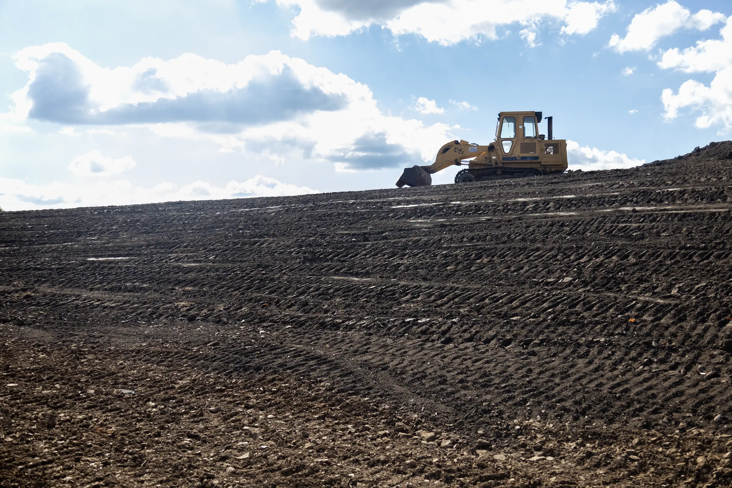 yellow bulldozer leveling dark soil on a sloped construction site under a partly cloudy sky