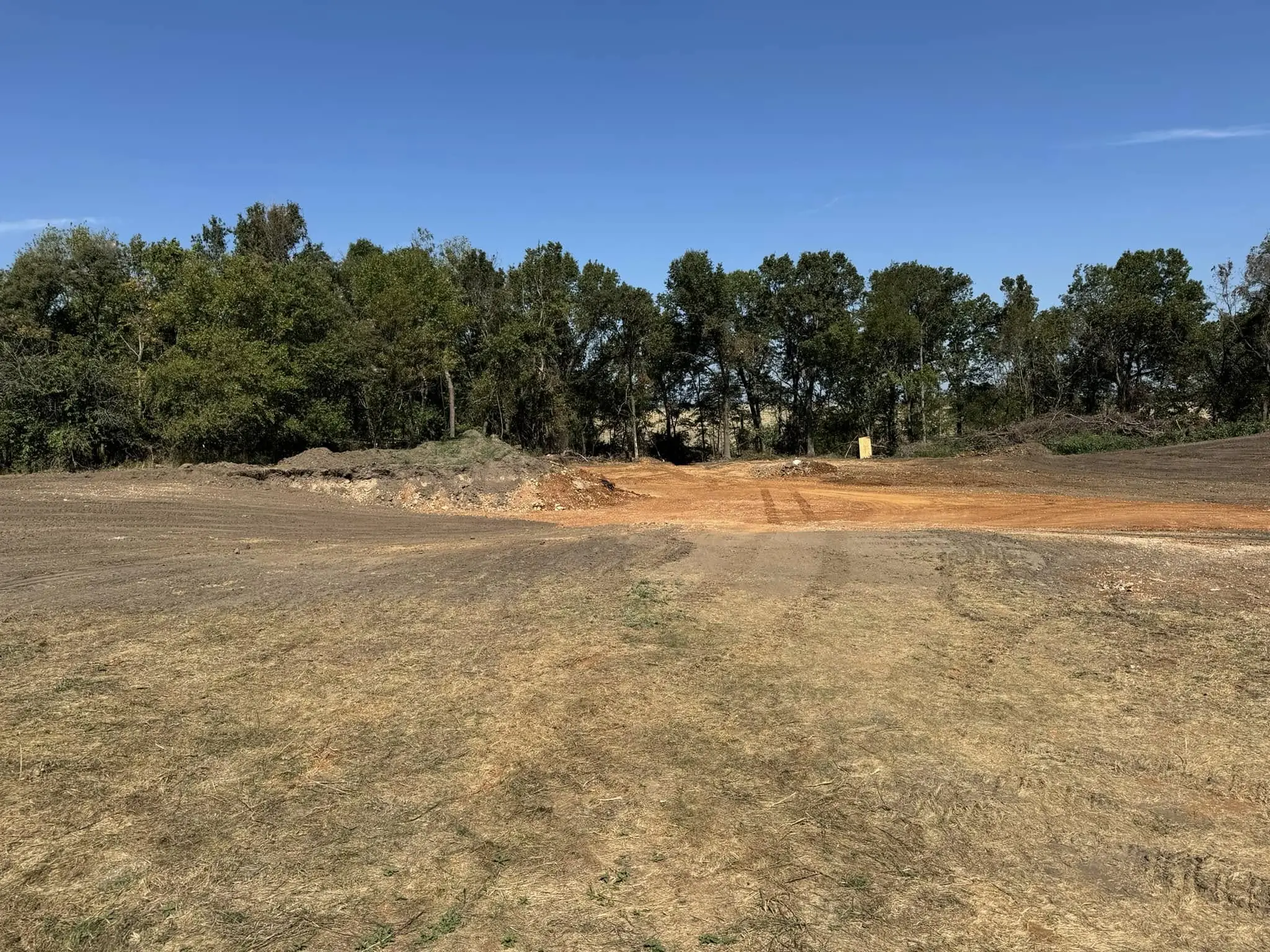 graded dirt field with leveled soil and small mounds of earth bordered by trees under a clear blue sky