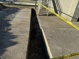 Narrow trench cut through concrete pavement beside a building marked off with yellow caution tape