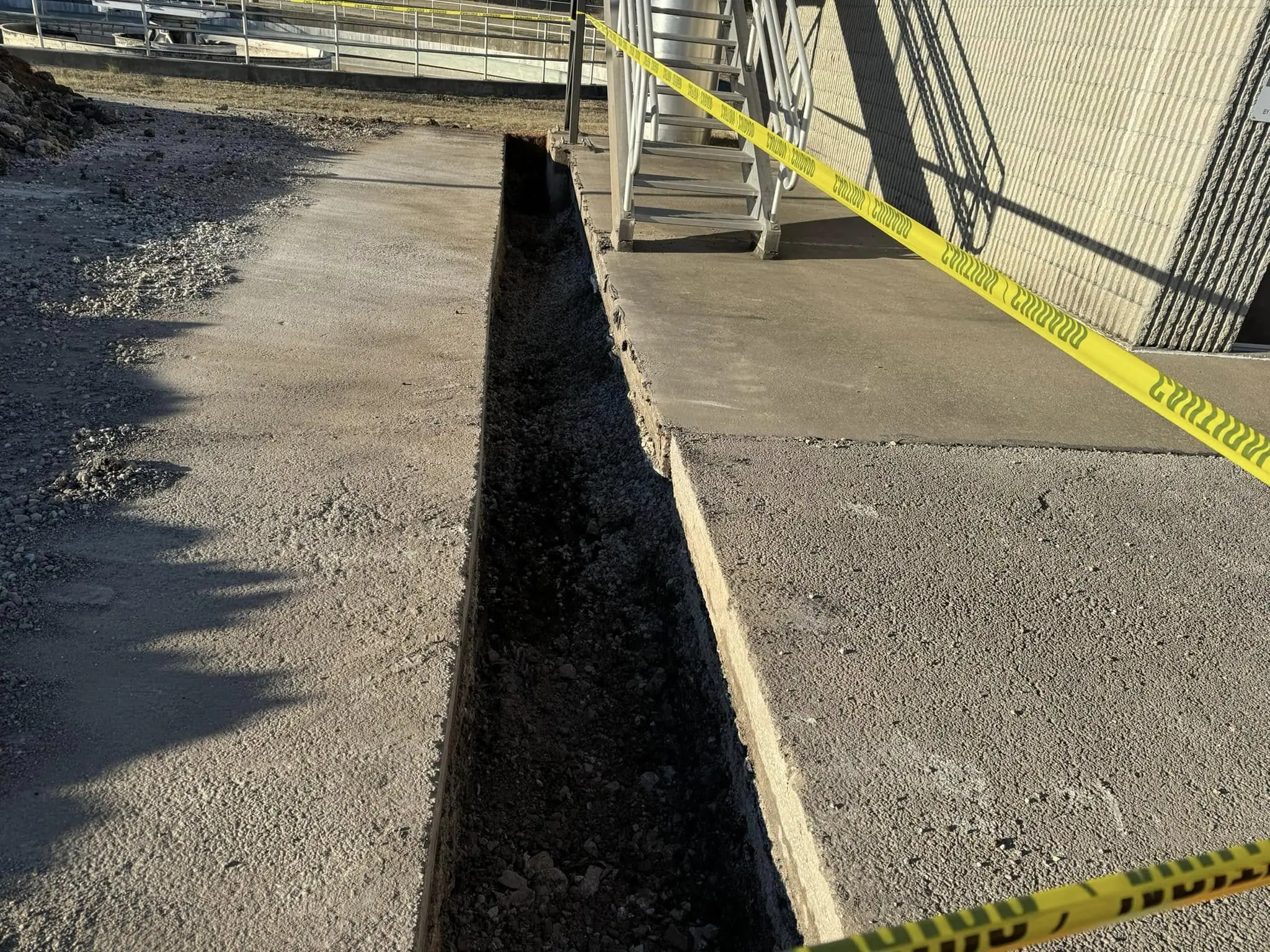 Narrow trench cut through concrete pavement beside a building marked off with yellow caution tape
