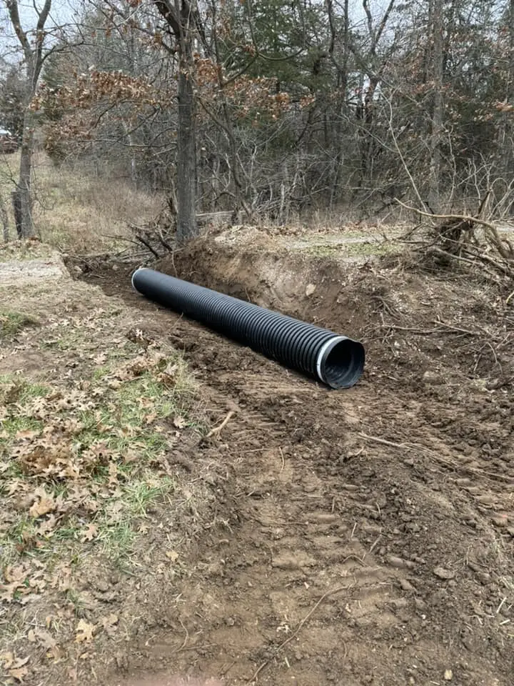 black corrugated plastic pipe positioned in a freshly dug soil trench beside a wooded area with sparse grass