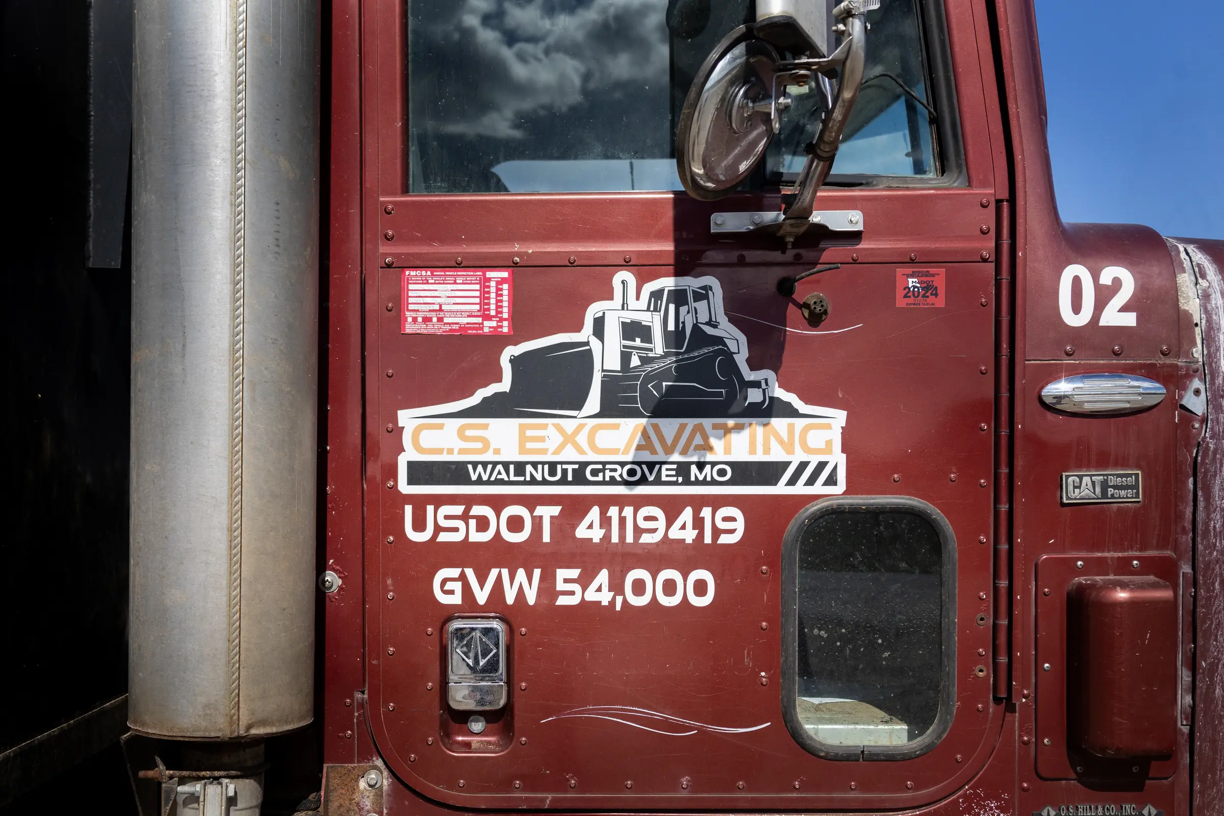 red dump truck door with excavation company markings and reflective metal exhaust stack under a partly cloudy sky