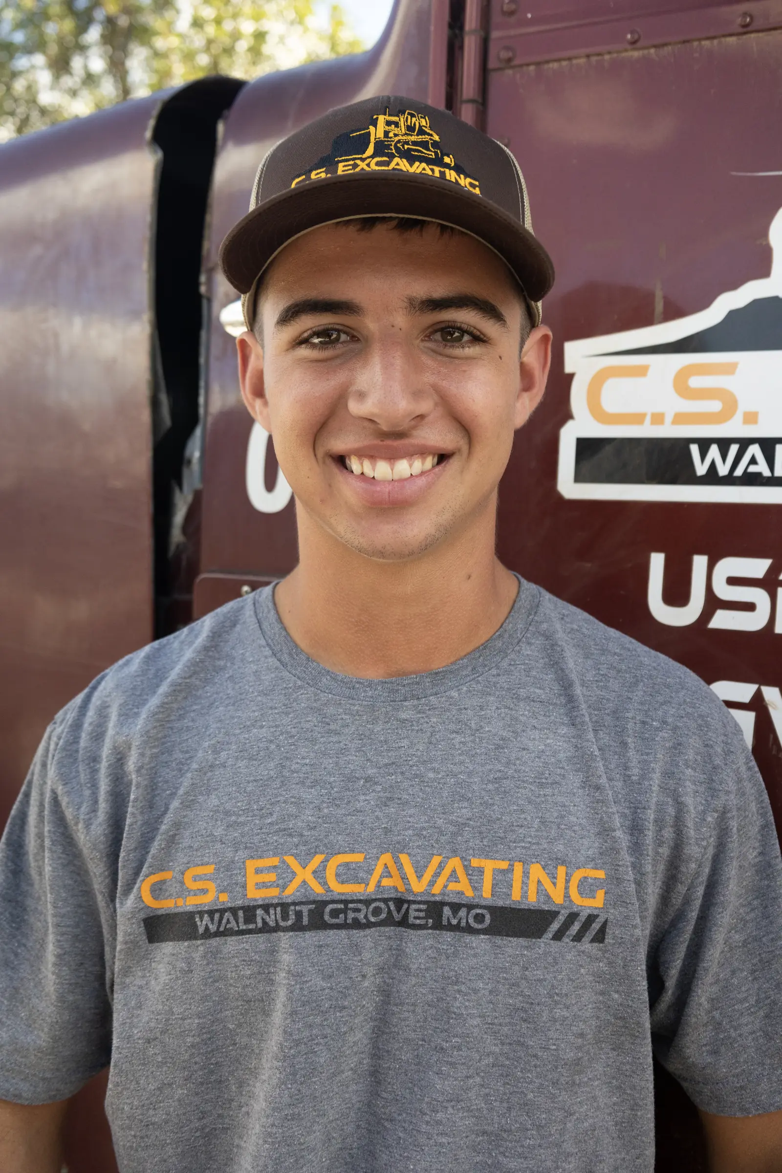 Worker wearing a gray excavation company shirt and hat standing beside a maroon work truck at a job site