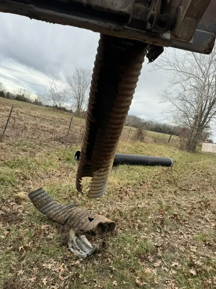 yellow excavator arm lifting a broken corrugated drain pipe over grassy ground with replacement pipe nearby