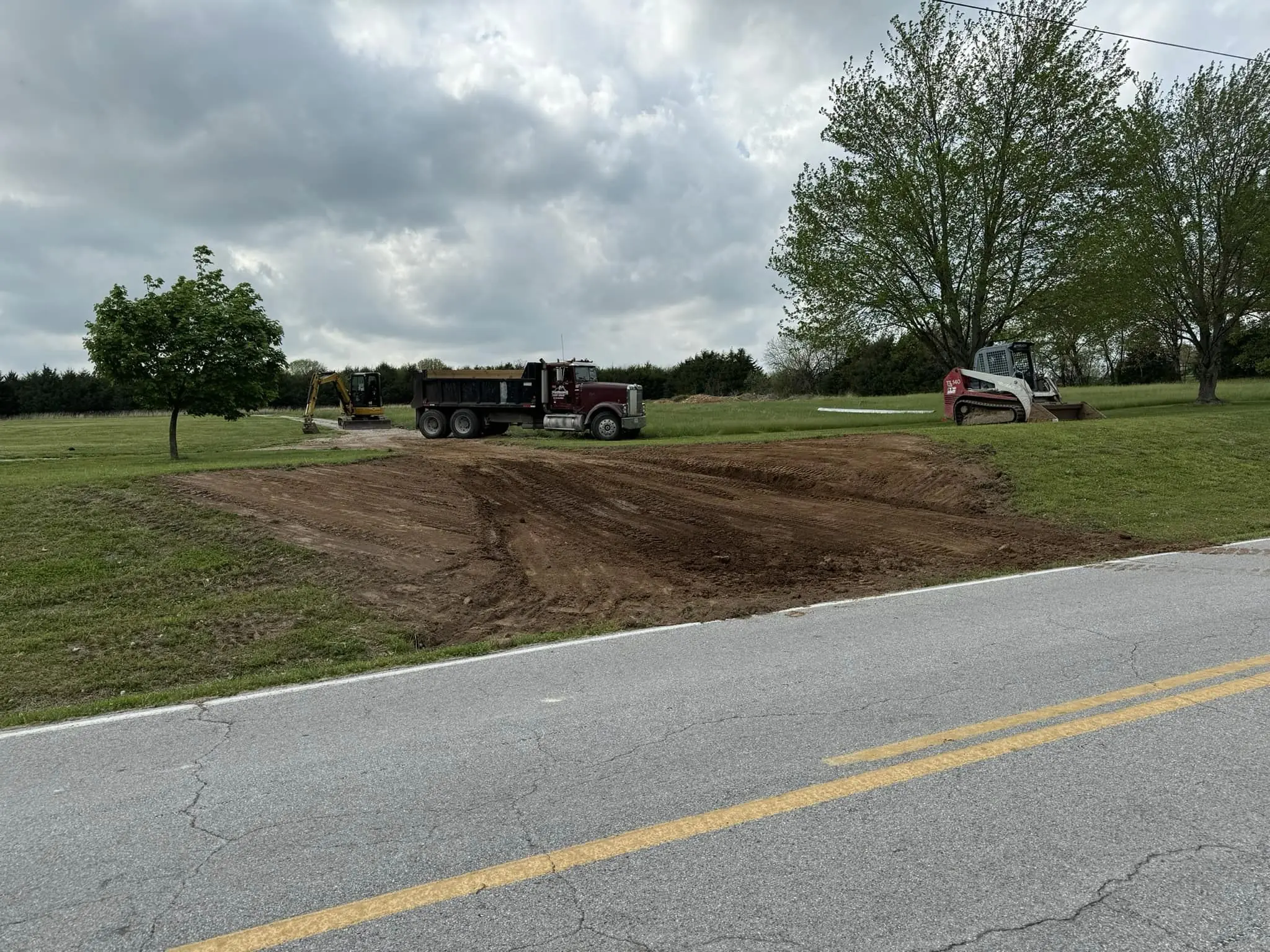 red skid steer and yellow excavator grading a dirt driveway entrance beside a dump truck on a grassy roadside