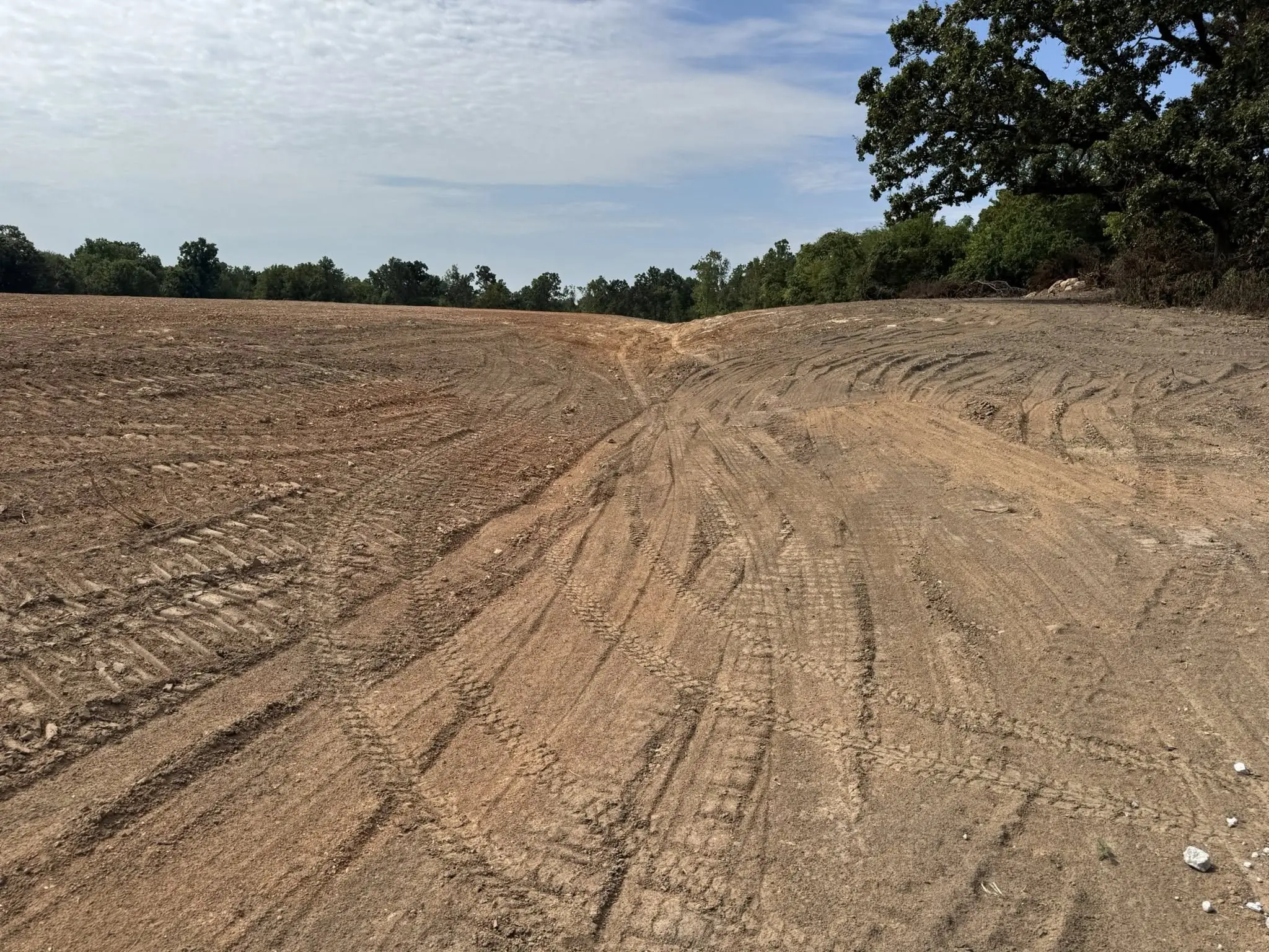 freshly graded dirt field with visible tire tracks beside a tree line under a partly cloudy sky