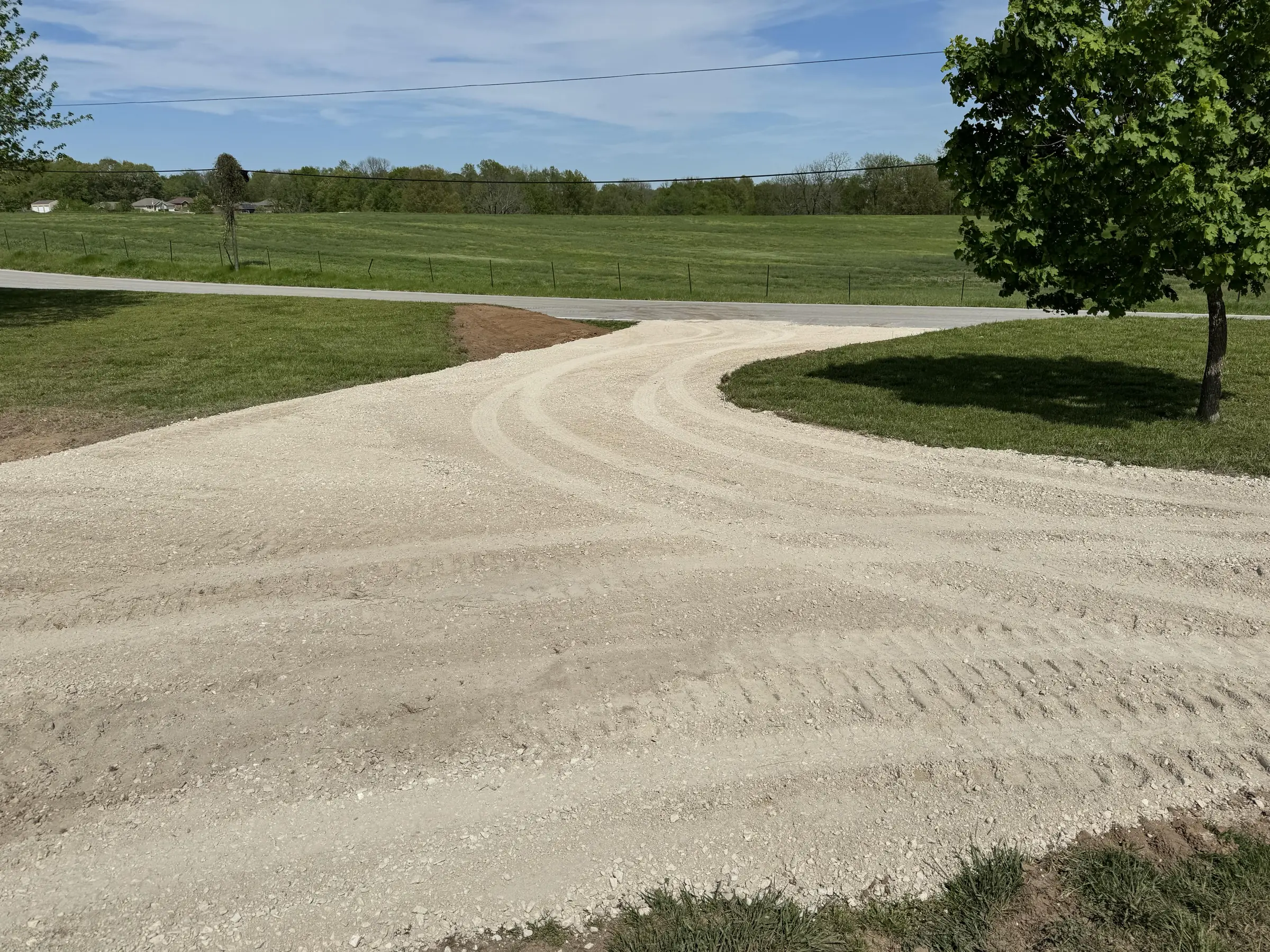 freshly leveled gravel driveway curving through a grassy yard with a tree and open field in the background