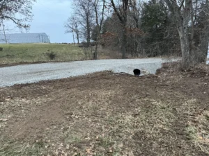 freshly laid gravel driveway with a metal culvert pipe beneath it beside trees and a grassy field