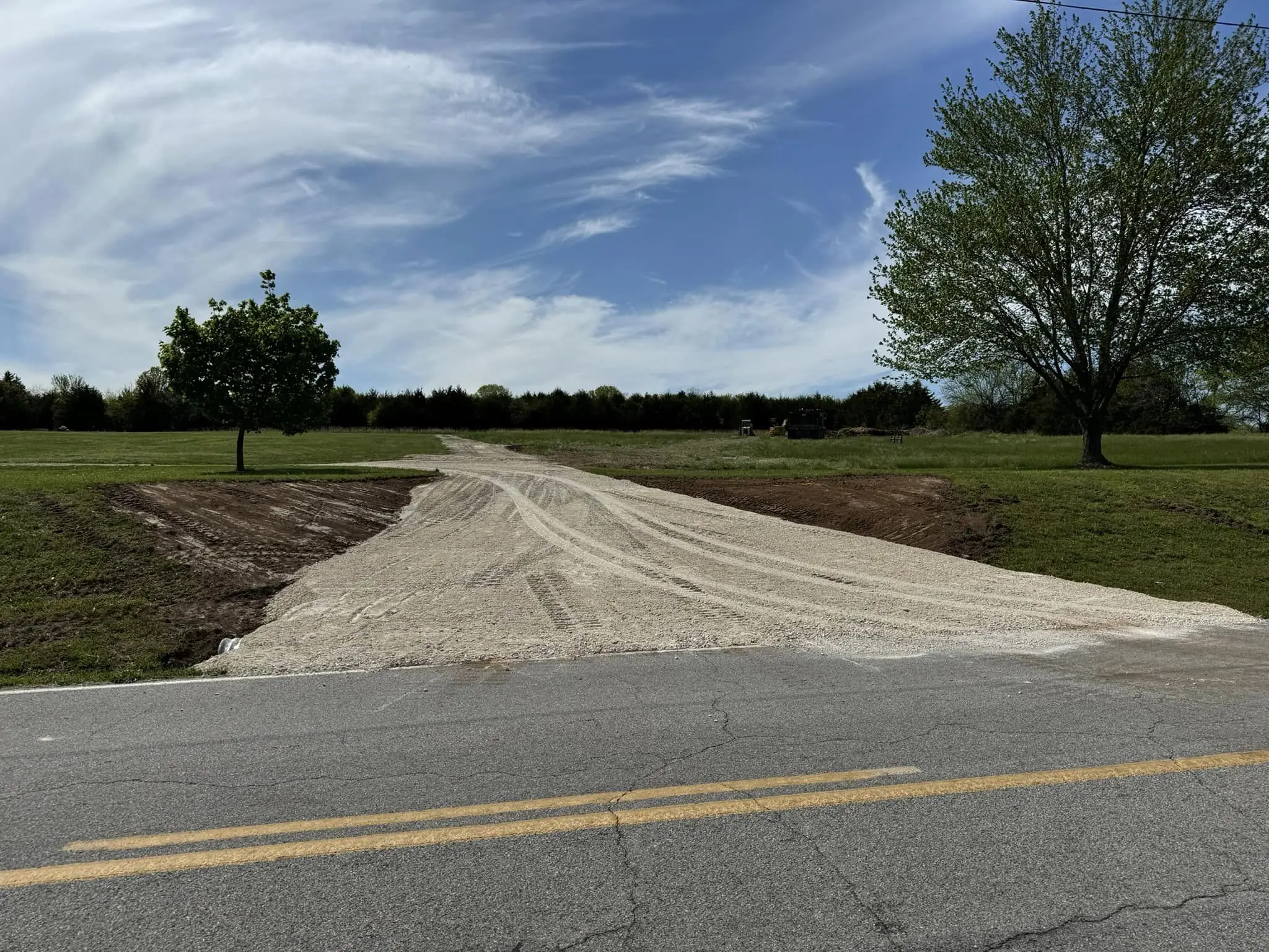 fresh gravel driveway graded from paved road through grassy field bordered by trees under a partly cloudy sky