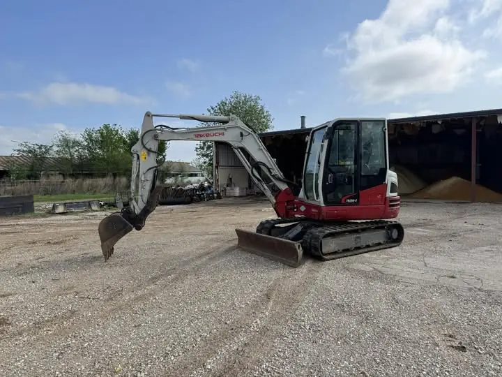 compact red and gray excavator parked on a gravel lot near open storage sheds and scattered construction materials