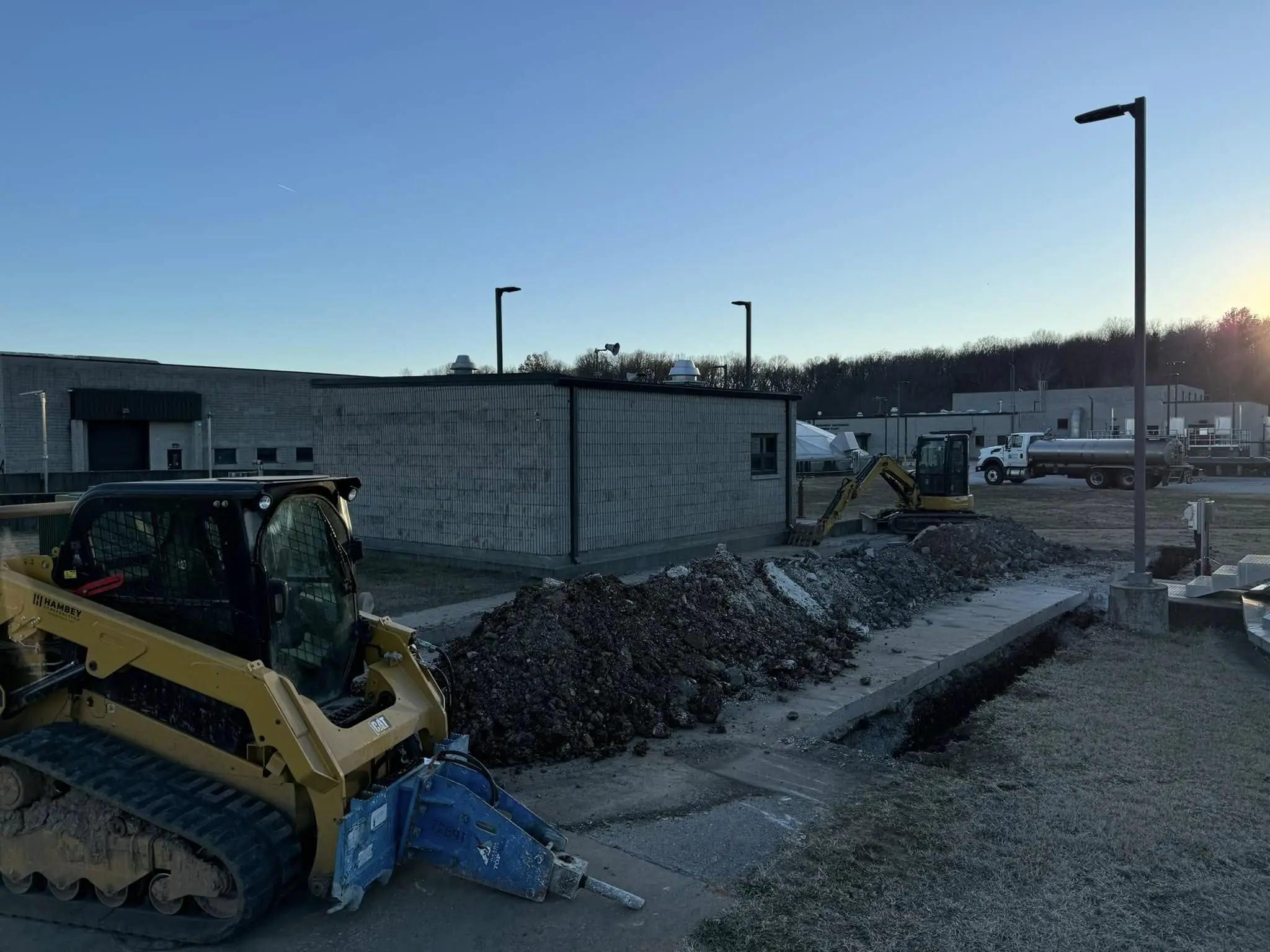 Yellow track loader and mini excavator digging along a dirt trench beside a concrete block building at sunrise