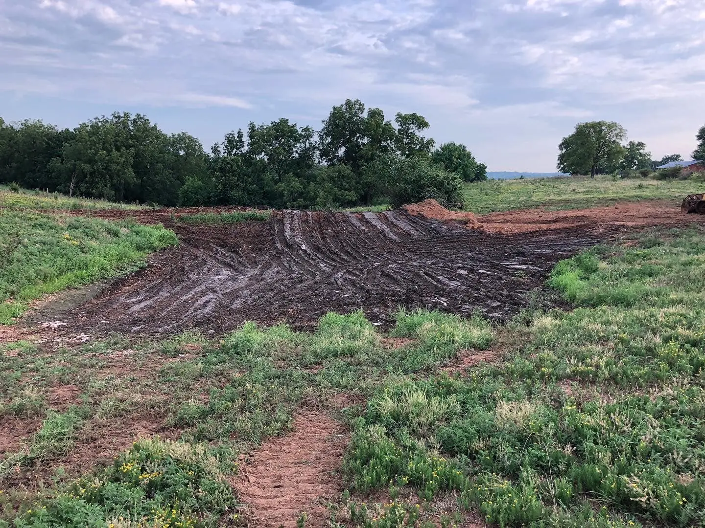 partially excavated dirt pond basin with fresh grading marks surrounded by grassy field and scattered trees