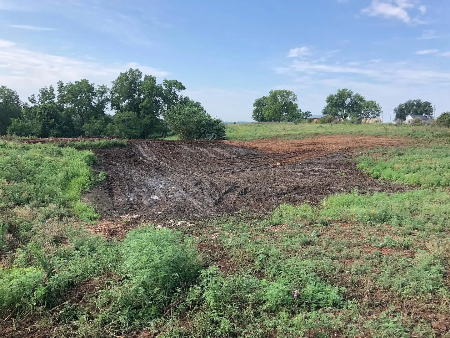 freshly excavated dirt basin surrounded by grassy field with trees in the background under a partly cloudy sky