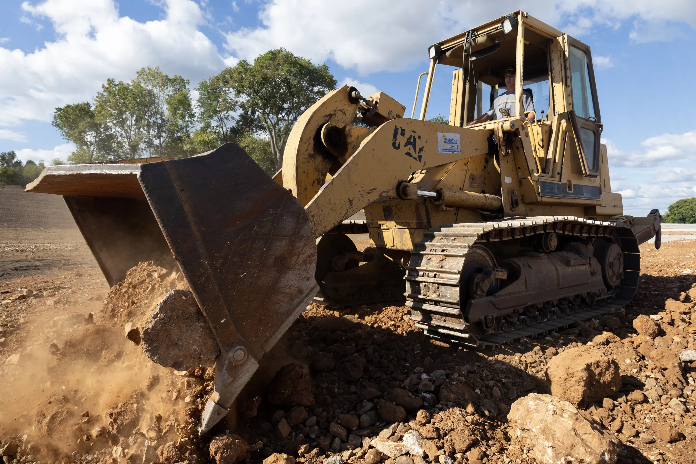 yellow track loader lifting loose soil and rocks on a dusty construction site with trees in the background