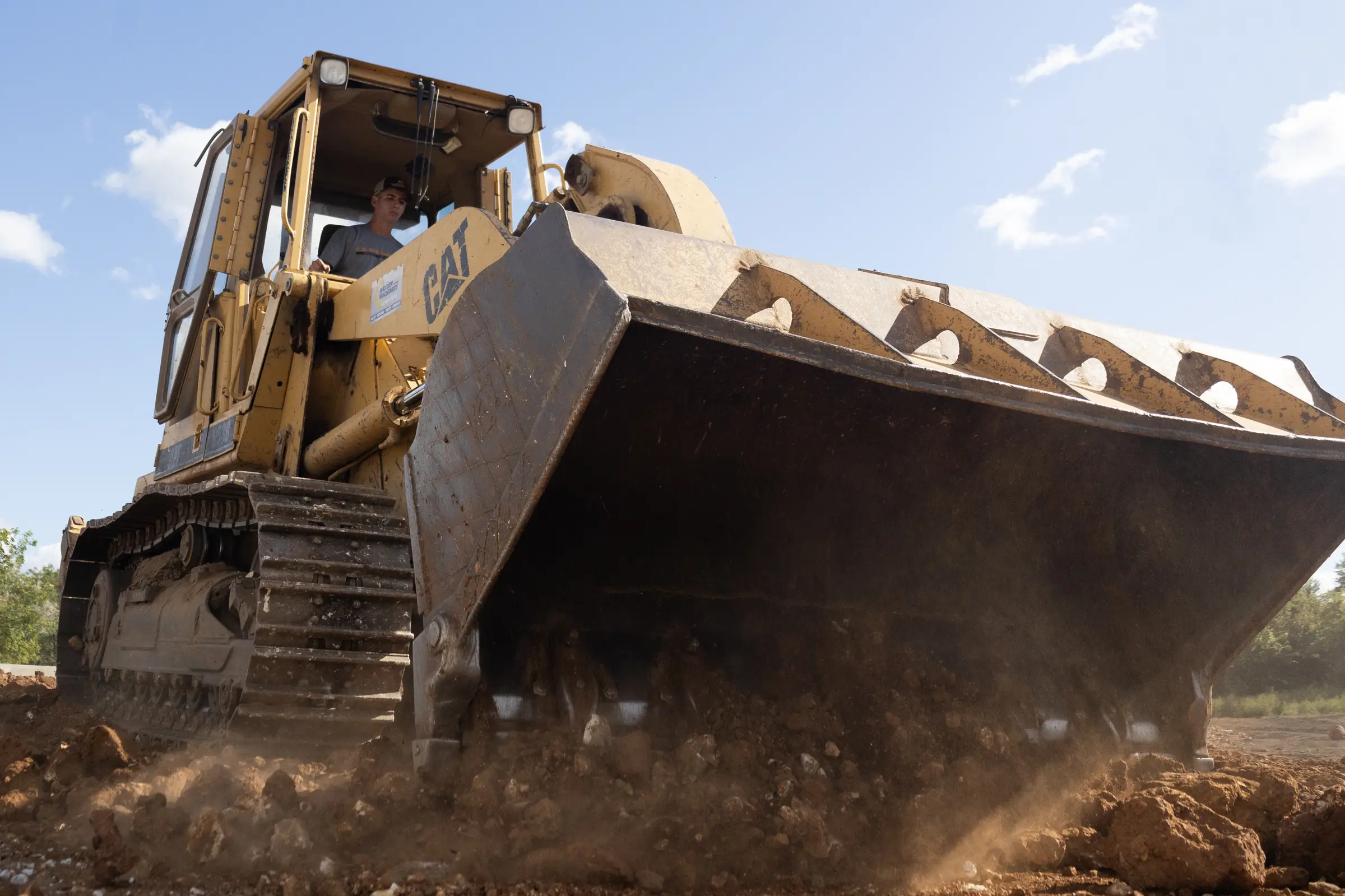 yellow track loader pushing loose soil and rocks across a dusty grading site under a clear blue sky