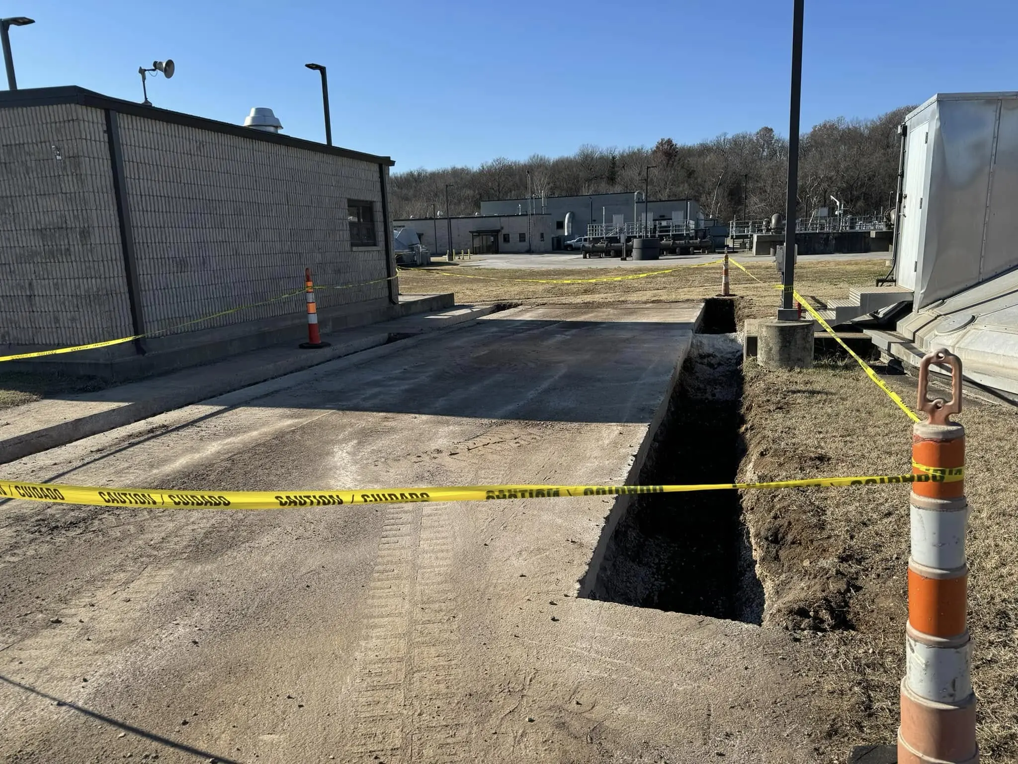 deep trench cut along paved area beside a block building with caution tape and traffic cones marking the site